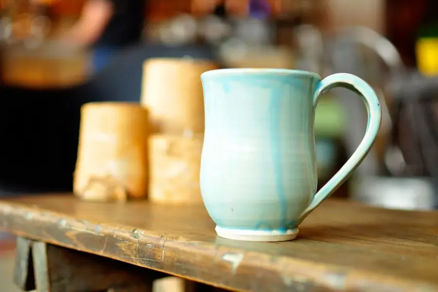 A pale turquoise glazed mug on a workshop bench beside unglazed clay pieces